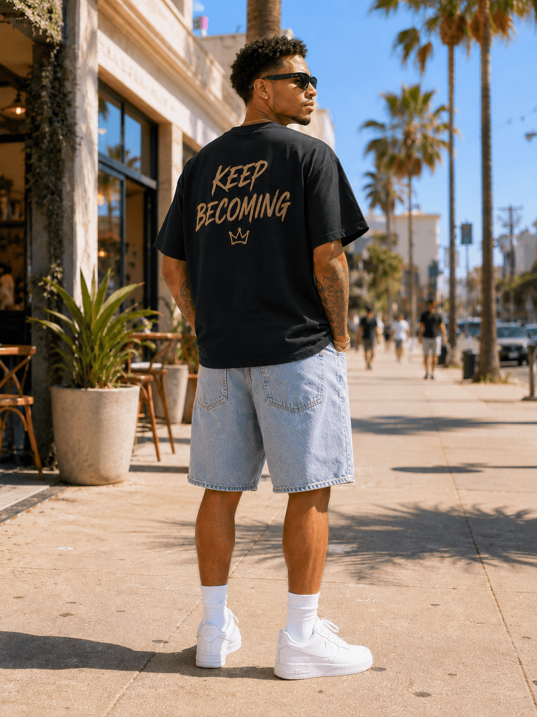Back of The Becoming Tee showing the Keep Becoming print and crown, on a palm-lined street